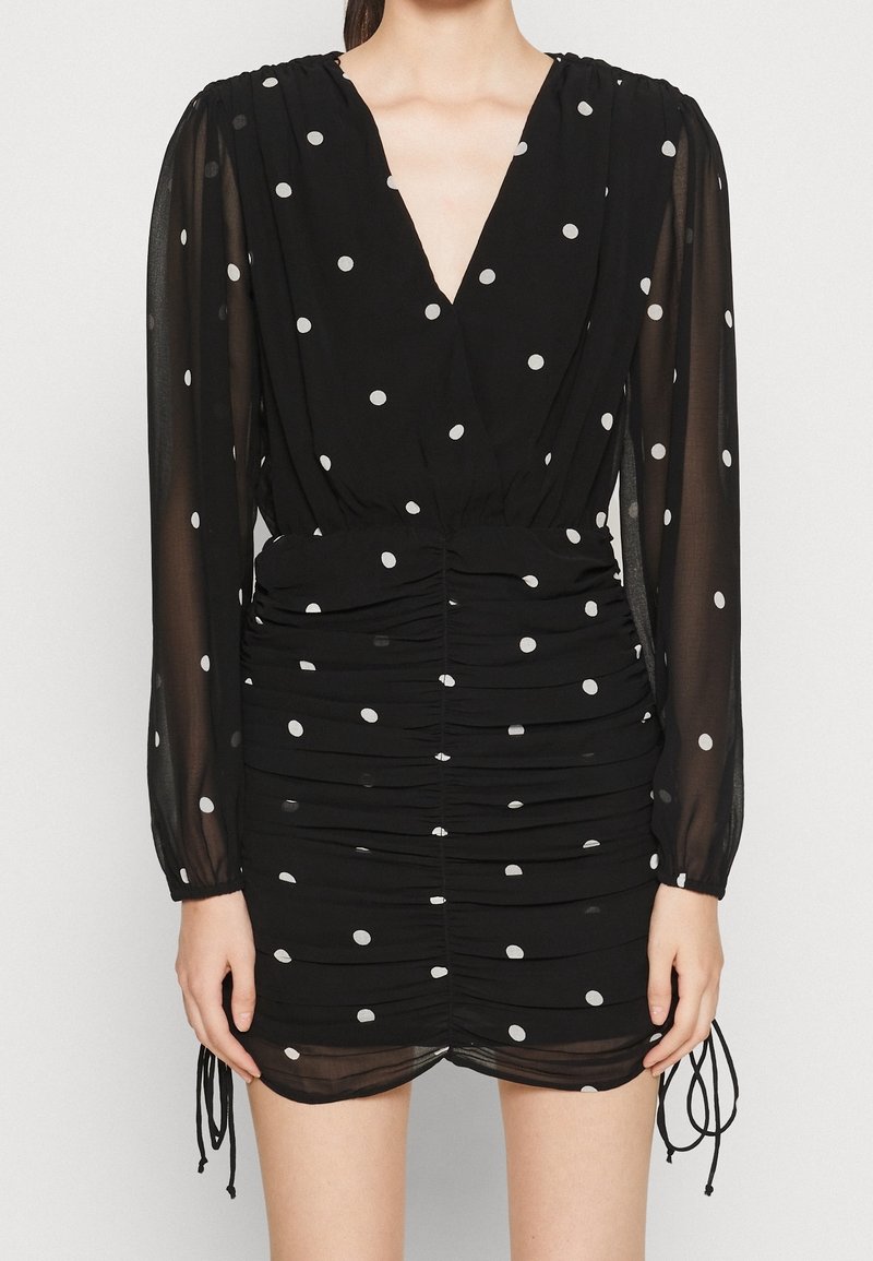 Svart klänning med djup V-ringning, långa genomskinliga ärmar, rynkade sidor och vita prickar. Inkluderar dragsko vid fållen.