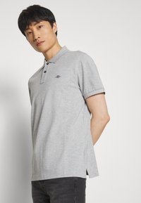 Grå polo-shirt med krave, korte ærmer og slidser i siderne. Sort knaplukning og et lille logo på venstre brystområde.