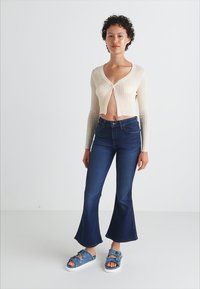 Geribbelde beige cropped cardigan, donkere flared jeans en blauwe denim slides met gespsdetails. Zachte texturen en aansluitende ontwerpelementen.