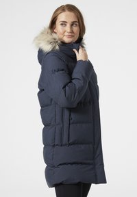 Námorněmodrá péřová bunda s kapucí lemovanou kožešinou, s vertikálními prošívanými částmi, předními kapsami a lemem sahajícím pod kolena.