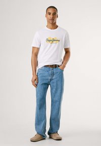 Balta trumpomis rankovėmis marškinėliai su mėlynu ir geltonu "Pepe Jeans London" logotipu, derinami su šviesiai mėlynomis tiesiomis džinsomis ir smėlio spalvos šlepetėmis.