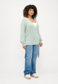 Μαλακή πράσινη, υφασμένη ζακέτα με μακριά μανίκια και πλάγιο δέσιμο, φορεμένη πάνω από λευκό τοπ, σε συνδυασμό με ανοιχτό μπλε τζιν και αθλητικά παπούτσια.