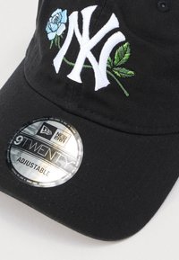 Čierna čiapka s vyšívaným logom NY v bielej farbe, zdobená modrou ružou a zelenými listami. Obsahuje strieborný nastaviteľný štítok na šiltu.
