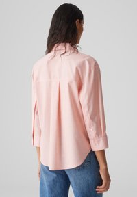 Hellrosa Langarmhemd mit Kragen, Knopfdetails und gefaltetem Rückenteil. Kombiniert mit blauen Denim-Jeans.