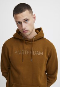 Sweatshirt à capuche marron avec "AMSTERDAM" imprimé en gris clair sur la poitrine. Doté de cordons de serrage et d'une poche kangourou. Texture douce.