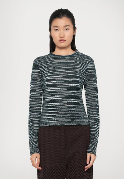 Langärmliges Oberteil in Marineblau und Teal, mit horizontalen wavy Mustern, runder Ausschnitt und strukturiertem Stoff. Kombiniert mit dunklen gemusterten Hosen.