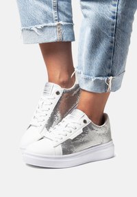 White sneakers with silver sequined panels, round toe, textured fabric, and flat white sole. Rolled-up light blue denim pants.