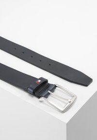 Cinturón de cuero negro con una hebilla de metal plateado. Presenta una hebilla rectangular y un pequeño detalle de logo cerca de la hebilla. Textura suave.