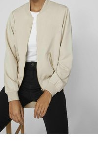 Beżowa kurtka bomberka z zapięciem na zamek, ściągaczami przy mankietach oraz dwoma bocznymi kieszeniami na zamek. Noszona na białej bluzce i w połączeniu z czarnymi spodniami.