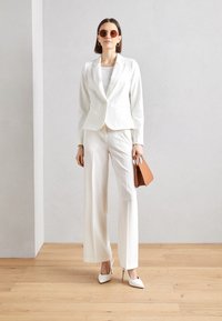 Weißer maßgeschneiderter Blazer und weit geschnittene Hose, kombiniert mit weißen High Heels und einer braunen Handtasche. Runde, braune Sonnenbrille vervollständigt den Look.