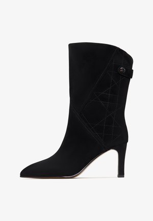 Sort ruskinds ankelstøvle med spids tå, slank hæl og kviltet geometrisk design på siden. Har en knapdetalje ved anklen.