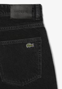 Los jeans de mezclilla negros cuentan con un bolsillo trasero que tiene un pequeño logo de cocodrilo en verde y rojo, y una etiqueta de cinturón de cuero negro suave marcada como "LACOSTE."