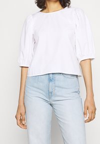 Bílý zkrácený top s nabíranými rukávy, kulatým výstřihem a hladkým bavlněným povrchem, doplněný o světle modré džíny s vysokým pasem.