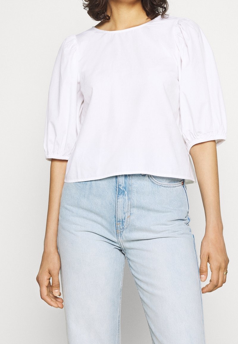 Bílý zkrácený top s nabíranými rukávy, kulatým výstřihem a hladkým bavlněným povrchem, doplněný o světle modré džíny s vysokým pasem.