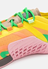 Sportovní obuv s vrchní částí z prodyšné síťoviny v žluté, oranžové a zelené barvě. Obsahuje zelené tkaničky, růžovou gumovou podrážku a strukturované detaily pro lepší přilnavost.