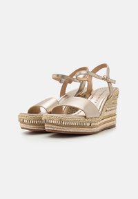 Sandales compensées dorées métalliques avec une plateforme en jute tressé, présentant une ouverture au niveau des orteils et des brides à la cheville. Texture lisse et détails de couture subtils.