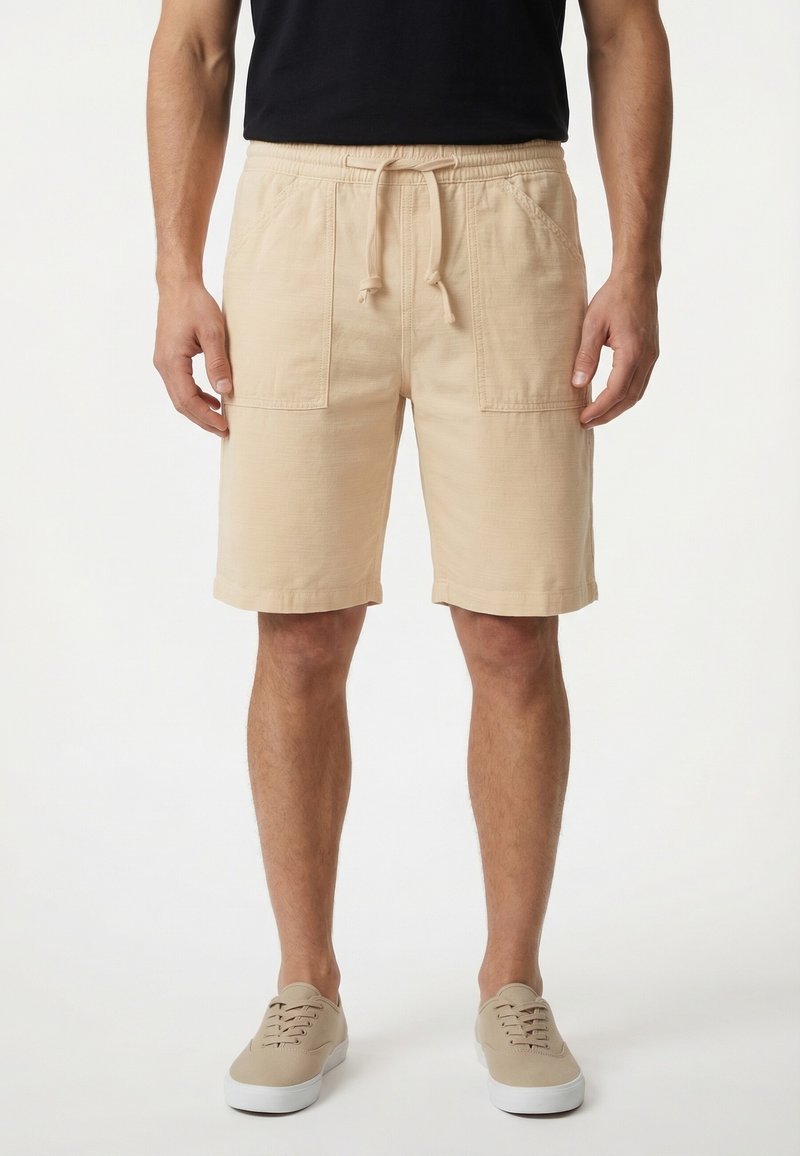 Homme portant un short beige à cordon de serrage arrivant aux genoux avec poches avant, un t-shirt noir et des baskets beige, debout devant un fond blanc.