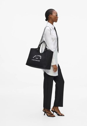 Femeie cu părul împletit poartă cămașă albă, cravată neagră, pantaloni negri, tocuri și poartă o geantă mare neagră Karl Lagerfeld pe umăr.