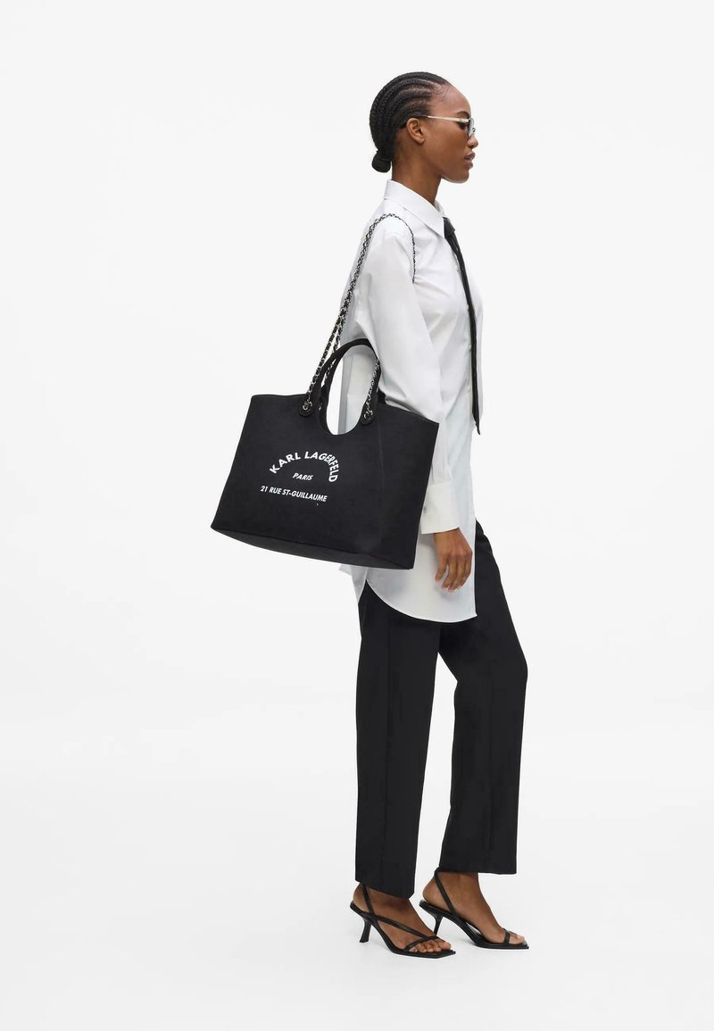 Femme aux cheveux tressés porte une chemise blanche, une cravate noire, un pantalon noir, des talons et porte un grand sac cabas noir Karl Lagerfeld sur l'épaule.