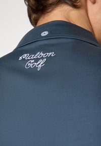 Marineblå golfskjorte med en struktureret overflade, der har en smal krave og en broderet "Malton Golf"-logo i hvid på ryggen.