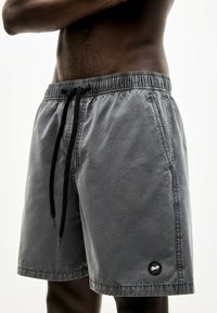 Graue Badehose mit verstellbarem Bund, glatter Textur, Seitentaschen und einem schwarzen, runden Logo am unteren Bein.