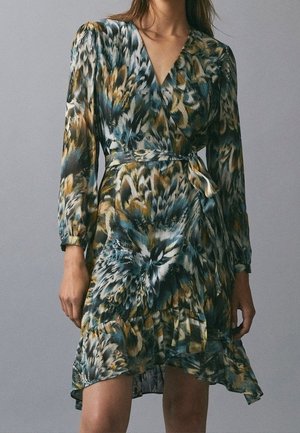 Femme portant une robe portefeuille à manches longues avec un motif abstrait de plumes bleu, vert et marron et une ceinture nouée à la taille.