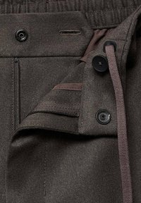 Tessuto dei pantaloni di colore marrone scuro con finitura testurizzata. Presenta una zip, bottoni e un cordoncino. Include un piccolo dettaglio con tasca interna.