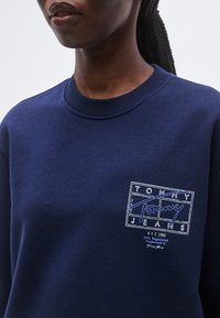 Teget plava majica dugih rukava s rebrastim izrezom, pamučnim materijalom i bijelom aplikacijom s uvezenim logotipom s plavim naglascima na prsima.