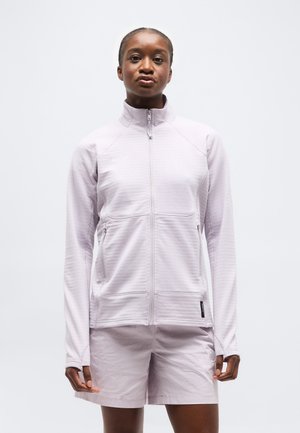 Nő világos lila cipzáras dzsekit és hozzáillő rövidnadrágot visel, egyszerű fehér háttér előtt áll.