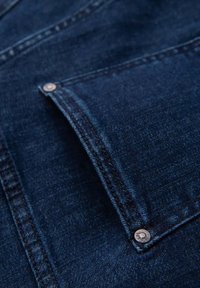 Détail d'une veste en denim avec un tissu bleu foncé, des coutures contrastées, une poche poitrine et des boutons en métal. Texture lisse et léger éclat.