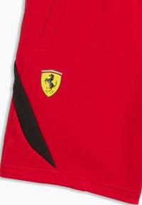 Pantalones cortos deportivos rojos con detalle triangular negro y logo amarillo de Ferrari que presenta un caballo encabritado y los colores de la bandera italiana.