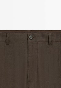 Gros plan de la ceinture d'un pantalon marron habillé avec fermeture par bouton et passants de ceinture, montrant la texture du tissu et des fines rayures verticales.