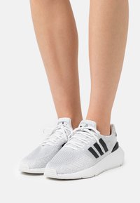 Sneaker bianche in maglia con accenti neri, dotate di una parte superiore testurizzata, suola ammortizzata e logo a tre strisce sul lato.