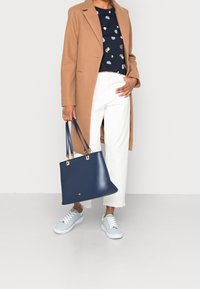 Bolso tote de cuero azul marino con doble asa, herrajes dorados y forma estructurada, combinado con un abrigo beige y pantalones cortos blancos.