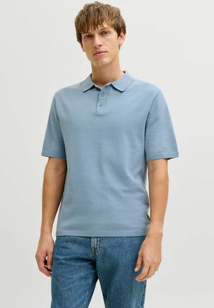 Jeune homme aux cheveux bruns courts portant un polo bleu clair à manches courtes et un jean bleu, debout devant un fond clair uni.