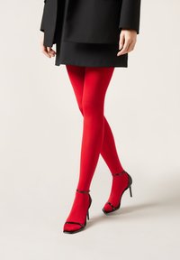 Červené neprůhledné punčocháče nošené s černými vysokými podpatky s lesklým akcentem a otevřenou špičkou, stylizované s černým kabátem.