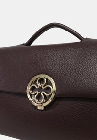 Sac en cuir bordeaux avec une surface texturée, une poignée arrondie et un fermoir circulaire en or avec un design floral complexe.