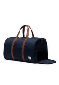Navy duffel bag yang terbuat dari kain dengan pegangan kulit coklat, dilengkapi dengan pembukaan ritsleting dan pintu samping untuk akses yang mudah.