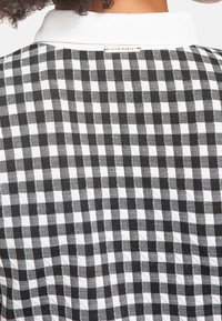 Mustavalkoinen pikkutäkkikuvioinen kangas, jossa on tekstuuri, valkoinen kaulus ja etiketti takana kauluksen sauman lähellä.