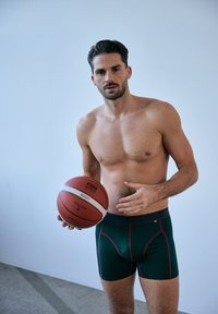 Gröna boxerkalsonger med en rosa midjeband, åtsittande design och slät textur. Den enskilda personen håller en basketboll och står framför en ljus bakgrund.