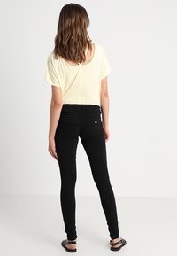 Helles gelbes, kurzärmeliges Oberteil mit weitem Ausschnitt, kombiniert mit schwarzen engen Jeans. Das Outfit wird durch flache schwarze Sandalen ergänzt.