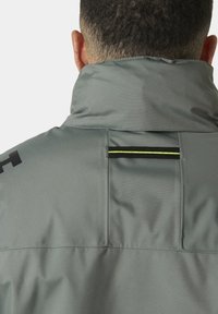 Grijze waterdichte jas met een hoge kraag, voorzien van een verticale zwarte streep en neon groene accenten. Textuur, duurzaam materiaal.