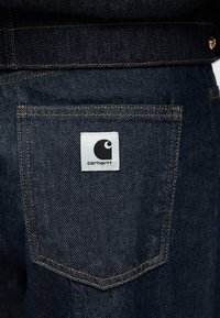 Σκούρο μπλε παντελόνι ντένιμ με πίσω τσέπη που φέρει λευκή ετικέτα με το λογότυπο Carhartt. Ξεχωριστή κίτρινη ραφή.