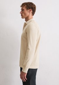 Beige Langarm-Polohemd aus weicher Stoffqualität, mit klassischem Kragen, Seitennähten und gerippten Bündchen, kombiniert mit dunklen Hosen.