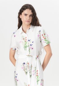 White floral dress featuring short sleeves, a button-up design, and a waist tie. Decorated with colorful flower patterns and green accents.