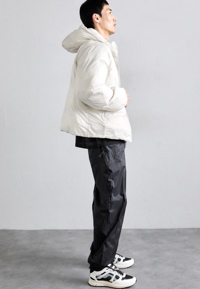 Dikke witte jas met een capuchon, gedragen over een zwart shirt. Zwarte, lichte broek met ritsen, en zwart-witte sportschoenen.
