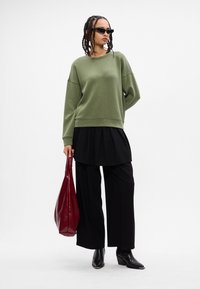 Sudadera verde oliva con corte oversized, combinada con una capa plisada negra y pantalones anchos negros. Bolso rojo brillante y botas negras de punta.