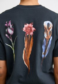Svart bomull t-skjorte med tre akvarellstil blomstertrykk i lilla, rød og blå med grønne og brune detaljer på baksiden.