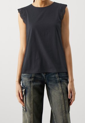 Kobieta ubrana w czarny top bez rękawów z koronkowym wykończeniem i wyblakłe niebieskie spodnie cargo, stojąca na jasnym, jednolitym tle.