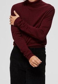Bordeauxroter Rollkragenpullover aus weichem Stoff, mit langen Ärmeln und gerippten Bündchen, kombiniert mit schwarzen Cordhosen.